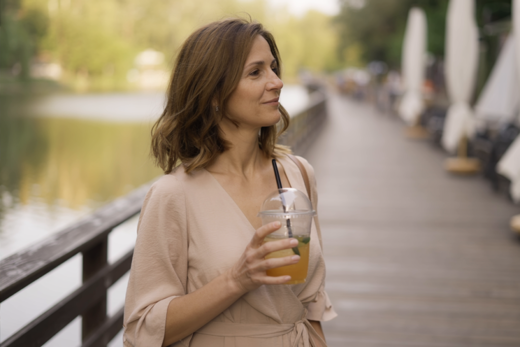 A woman in her 40s walks along a lakeside boardwalk, holding a plastic cup of iced lemon tea, wearing a soft wrap dress and looking peacefully ahead.