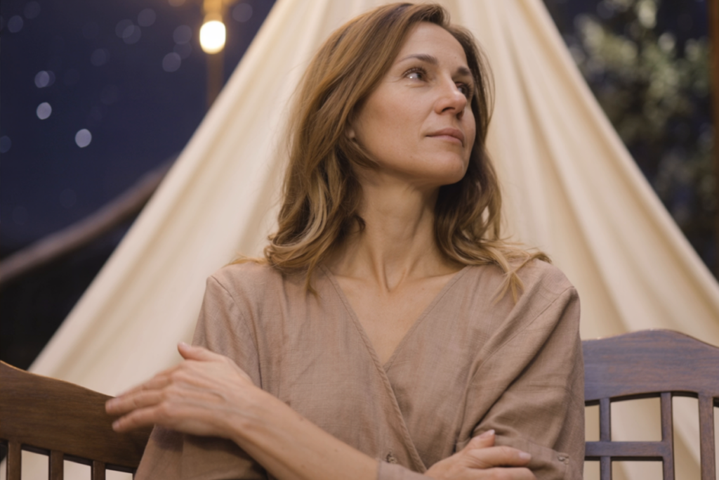 Elegant woman in soft lighting gazes upward at night, wrapped in beige, reflecting quiet sadness and longing.